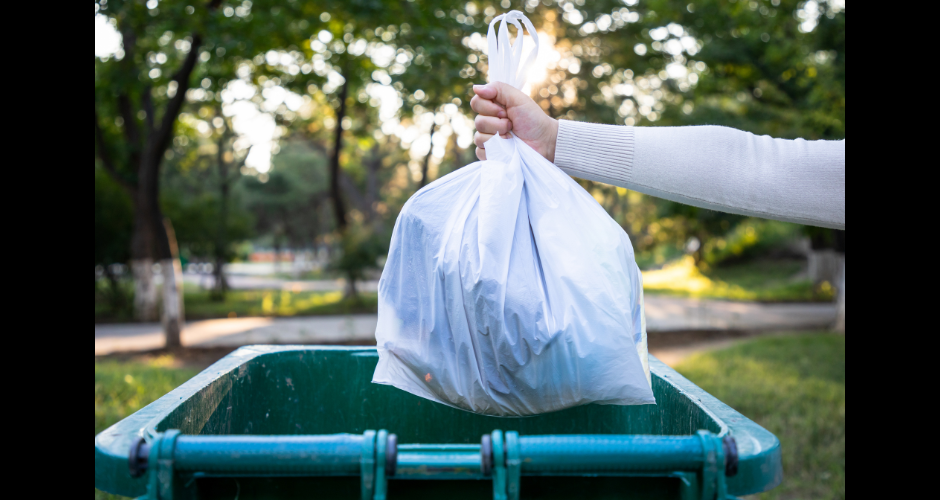 An arm drops a bag of trash into a trash receptacle. It's a blue receptacle.