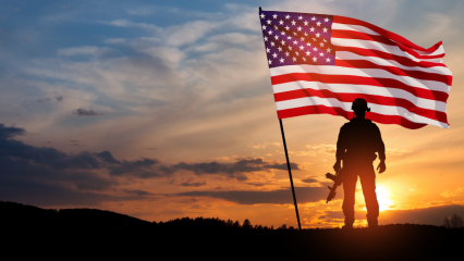 A soldier stands next to an American flag as the sun rises on the horizon.