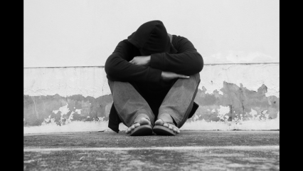 A homeless man in a black hoodie leans against a white wall.