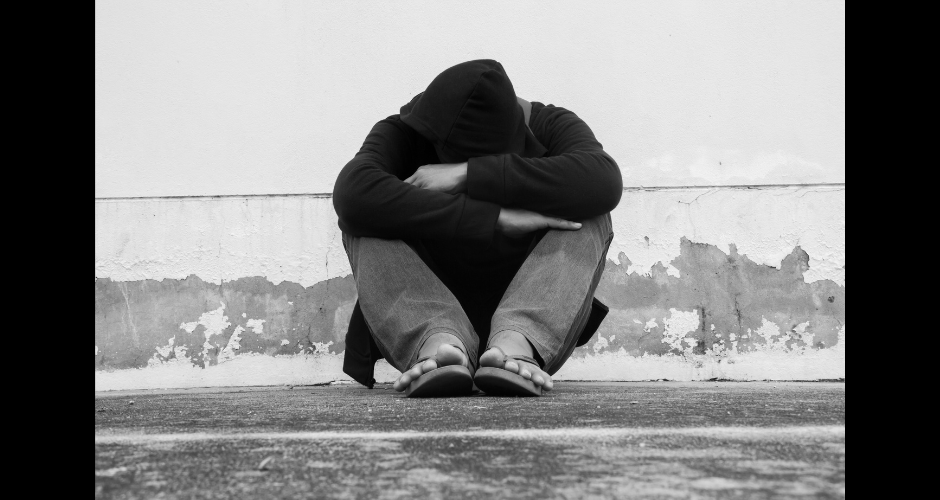 A homeless man in a black hoodie leans against a white wall.