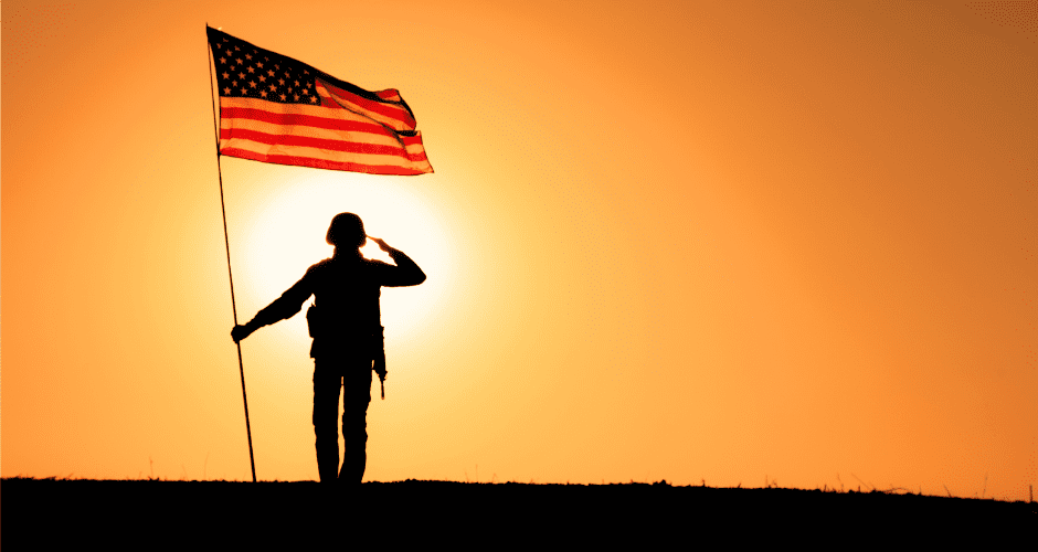 A soldier holding an American flag on a flagpole salutes will staring directly into the sunset.