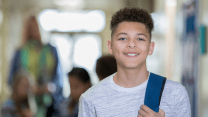 A smiling student clad in a white t-shirt stands with a backpack slung over his shoulder in the hallway of a high school
