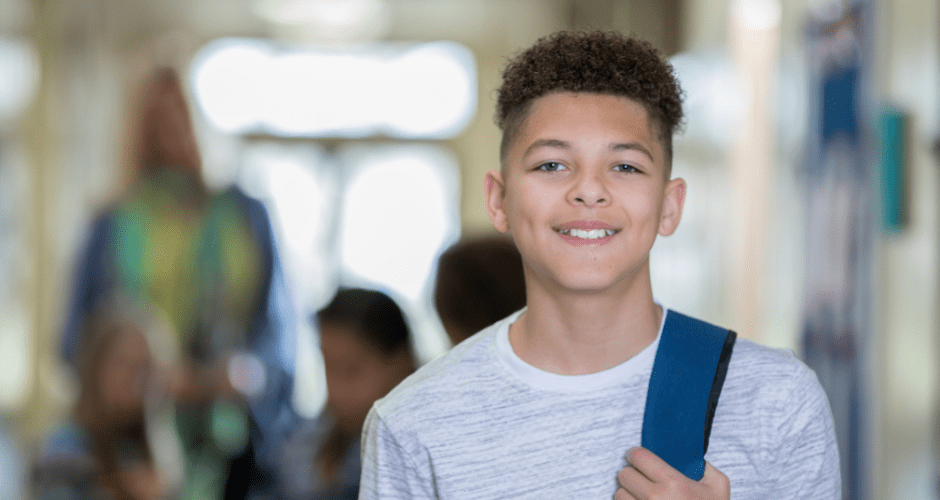 A smiling student clad in a white t-shirt stands with a backpack slung over his shoulder in the hallway of a high school
