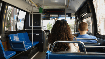 Two students sit on an otherwise empty bus. One is a boy with short hair and a blue shirt; the other is a girl with long brown hair and a white shirt. The bus has blue seats.
