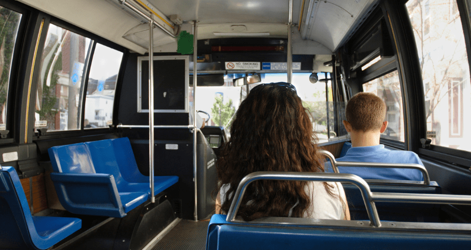 Two students sit on an otherwise empty bus. One is a boy with short hair and a blue shirt; the other is a girl with long brown hair and a white shirt. The bus has blue seats.