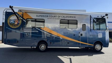 Side view of a blue and gold mobile unit branded “Superior Court of California, County of San Bernardino – Justice in Motion,” parked outdoors, featuring the county seal, mountain graphic, service windows, and a fold-out awning on a sunny day.