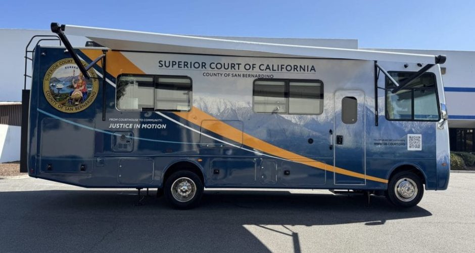 Side view of a blue and gold mobile unit branded “Superior Court of California, County of San Bernardino – Justice in Motion,” parked outdoors, featuring the county seal, mountain graphic, service windows, and a fold-out awning on a sunny day.