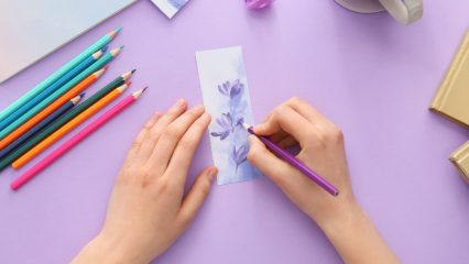 Hands drawing a lavender flower with a purple pencil on a bookmark, surrounded by colored pencils and a cup of tea.