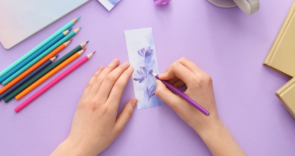 Hands drawing a lavender flower with a purple pencil on a bookmark, surrounded by colored pencils and a cup of tea.