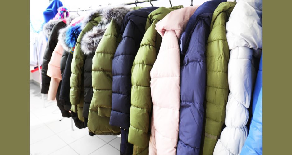A row of winter coats in various colors and styles hangs neatly on a clothing rack indoors, including puffy jackets with hoods and faux-fur trim, ready for donation to help keep people warm during cold weather.