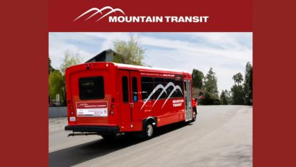 Red Mountain Transit shuttle bus with white mountain logo driving up a wide paved road on a sunny day, with trees and a building in the background.