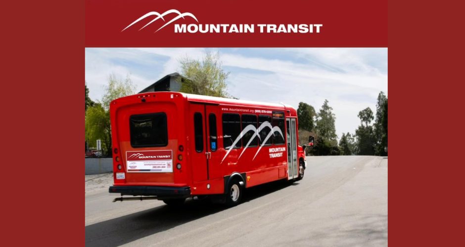 Red Mountain Transit shuttle bus with white mountain logo driving up a wide paved road on a sunny day, with trees and a building in the background.