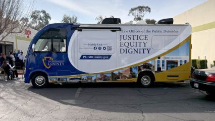 A blue and gold San Bernardino County Public Defender Mobile Defense Program vehicle is parked in a shopping center parking lot. The side of the mobile unit displays the words “Law Offices of the Public Defender” and “Justice, Equity, Dignity.” Several people are seated nearby at tables, with buildings and trees visible in the background under a clear sky.