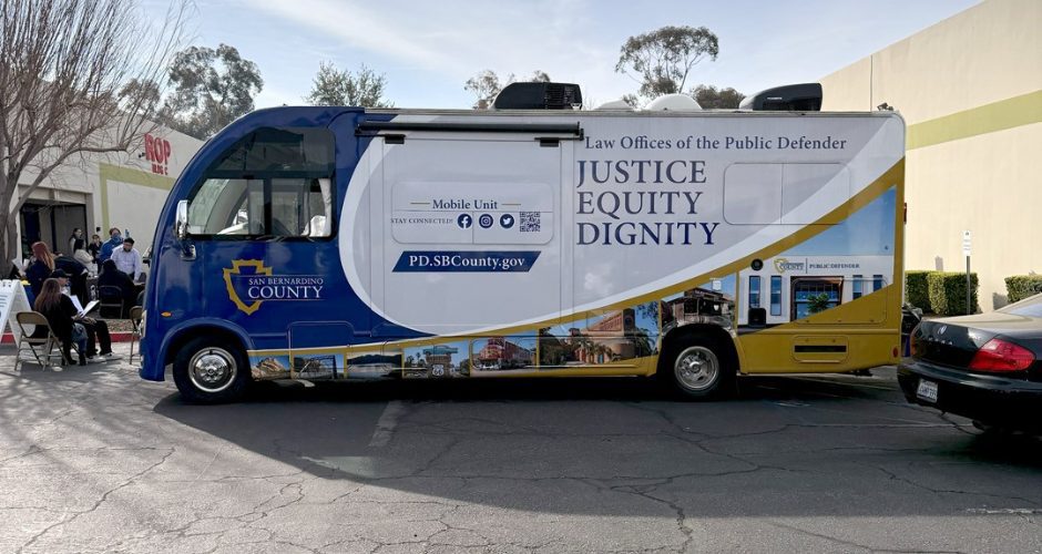 A blue and gold San Bernardino County Public Defender Mobile Defense Program vehicle is parked in a shopping center parking lot. The side of the mobile unit displays the words “Law Offices of the Public Defender” and “Justice, Equity, Dignity.” Several people are seated nearby at tables, with buildings and trees visible in the background under a clear sky.