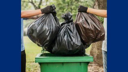 Two people wearing gloves lift large black trash bags into a green waste container outdoors, representing community cleanup and proper waste disposal.