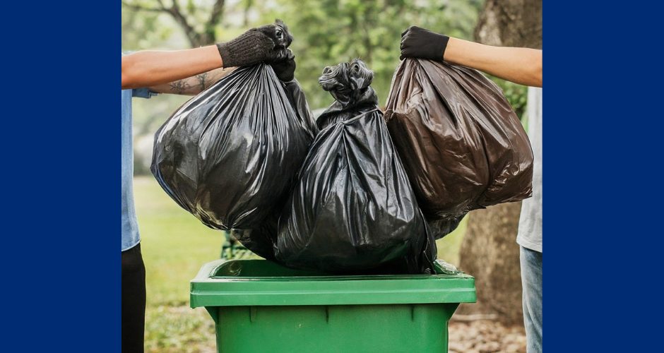 Two people wearing gloves lift large black trash bags into a green waste container outdoors, representing community cleanup and proper waste disposal.