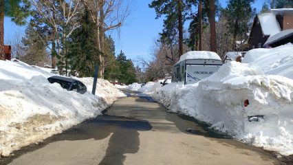 Snow-lined mountain neighborhood street with tall pine trees and cabins, where high plowed snowbanks tower on both sides of a narrow, partially wet roadway. A car is partially buried in snow on the left, and a white RV trailer labeled “Rogue” is parked on the right, surrounded by deep snow under a clear blue sky.