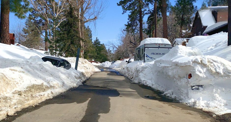 Snow-lined mountain neighborhood street with tall pine trees and cabins, where high plowed snowbanks tower on both sides of a narrow, partially wet roadway. A car is partially buried in snow on the left, and a white RV trailer labeled “Rogue” is parked on the right, surrounded by deep snow under a clear blue sky.