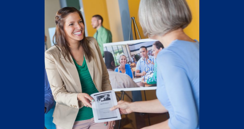 A woman at a job fair smiles as she hands a flyer to another attendee, with a recruitment display and people networking in the background.