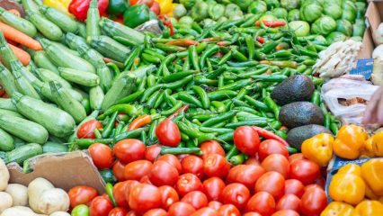 A colorful display of fresh produce at a farmers market, including red tomatoes, green chili peppers, zucchini, avocados, bell peppers, Brussels sprouts, and other vegetables arranged in rows on a table.