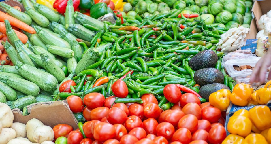 A colorful display of fresh produce at a farmers market, including red tomatoes, green chili peppers, zucchini, avocados, bell peppers, Brussels sprouts, and other vegetables arranged in rows on a table.