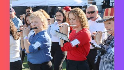 A dozen or so piccolo players stand next to each other, playing their instruments in an outdoor setting.