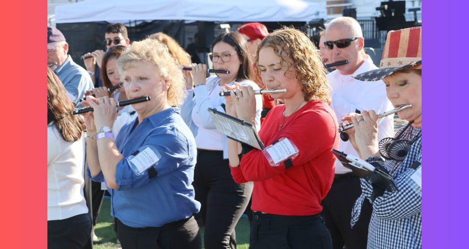 A dozen or so piccolo players stand next to each other, playing their instruments in an outdoor setting.
