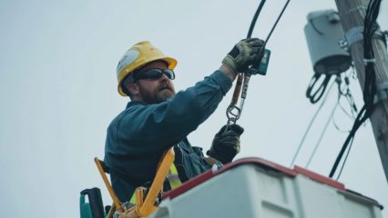 A utility worker wearing a yellow hard hat, safety gloves, and sunglasses works from the bucket of a utility truck while connecting or inspecting electrical equipment on a wooden utility pole. Power lines and utility hardware are visible against an overcast sky.