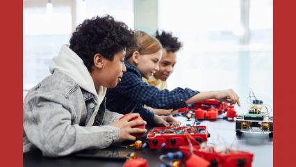 Three smiling kids work on a science-related project spread out on a table in front of them. One kid is wearing a hoodie.