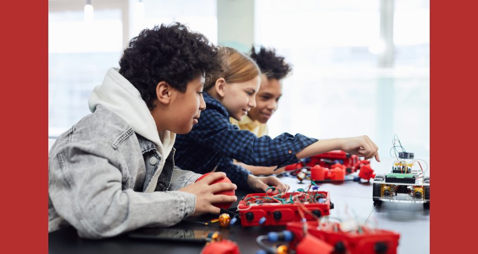 Three smiling kids work on a science-related project spread out on a table in front of them. One kid is wearing a hoodie.