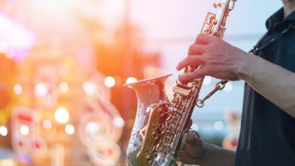 A saxophonist plays on an outdoor stage with what appears to be a drum set blurred in the background.