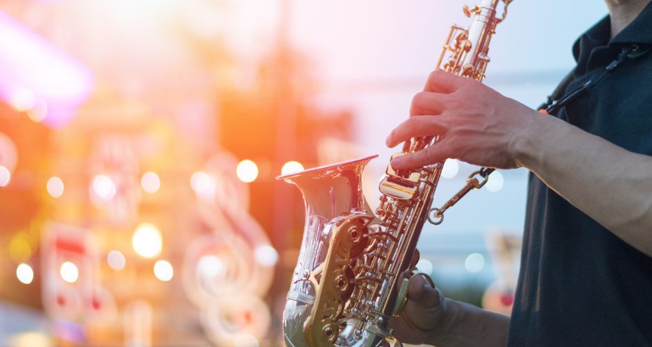 A saxophonist plays on an outdoor stage with what appears to be a drum set blurred in the background.