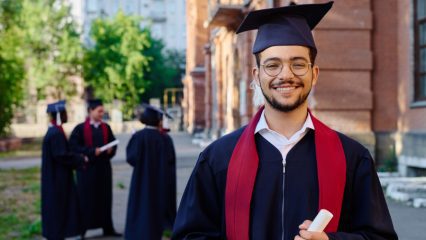 A smiling graduate wearing a black cap and gown with a red stole stands outdoors holding a rolled diploma. The graduate is in sharp focus in the foreground, while several other graduates in caps and gowns converse in the background near a campus building and trees, suggesting a college or graduation ceremony setting.