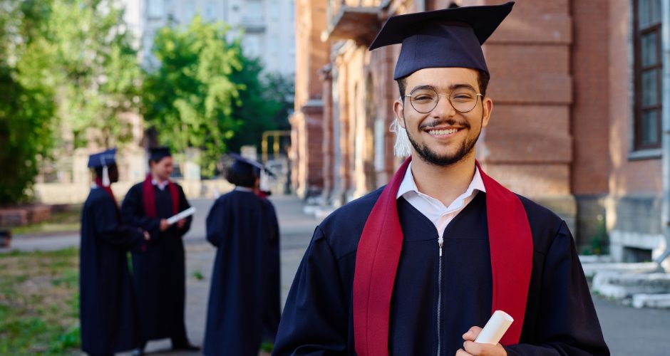 A smiling graduate wearing a black cap and gown with a red stole stands outdoors holding a rolled diploma. The graduate is in sharp focus in the foreground, while several other graduates in caps and gowns converse in the background near a campus building and trees, suggesting a college or graduation ceremony setting.