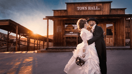 A newly married couple wearing traditional wedding clothing embrace in front of the Town Hall at Calico Ghost Town in Yermo, California as the sun slowly sets in the background.