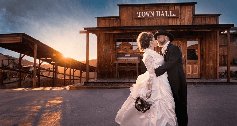 A newly married couple wearing traditional wedding clothing embrace in front of the Town Hall at Calico Ghost Town in Yermo, California as the sun slowly sets in the background.