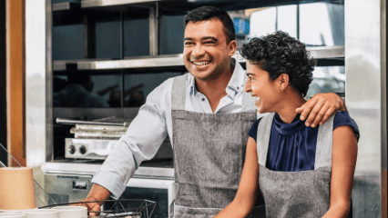 wo smiling small business owners wearing gray aprons stand together behind the counter in a commercial kitchen, with one person’s arm around the other.