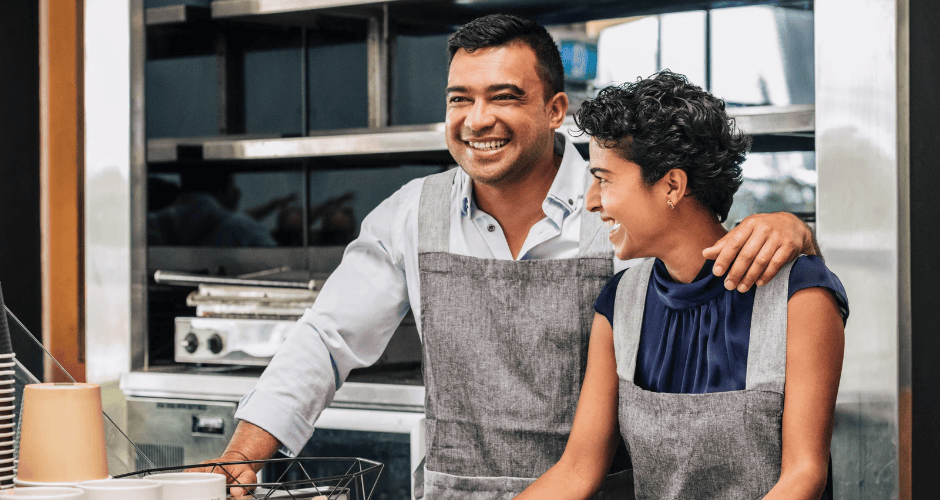 wo smiling small business owners wearing gray aprons stand together behind the counter in a commercial kitchen, with one person’s arm around the other.