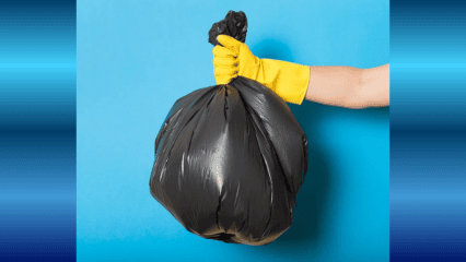 A single black trash bag is held by a yellow gloved hand against a light blue background.