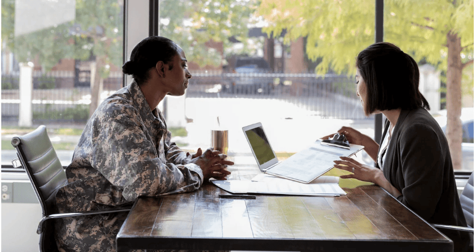 A woman in military uniform meets with a professional career counselor across a wooden table. The counselor reviews documents while the service member listens attentively. A laptop and paperwork sit between them in a bright office with large windows overlooking trees.